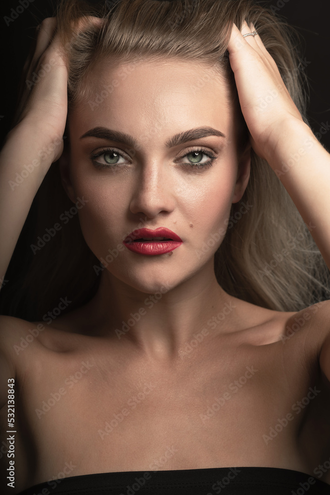 Fototapeta premium Studio portrait of beautiful blonde woman with long hair, green eyes and red lipstick looking to camera. Model holding hands over head and brushing hair with fingers. Toned image in beige color