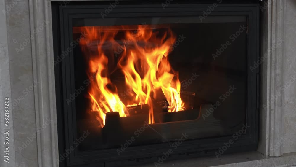 Stylish cast iron fireplace with fire in home interior lined with marble stone. Modern stove to decorate and heat house with the fire burning wood. Orange tongues of flame dance behind fireproof glass Stylish cast iron fireplace with fire in home interior lined with marble stone. Modern stove to decorate and heat house with the fire burning wood. Orange tongues of flame dance behind fireproof glass