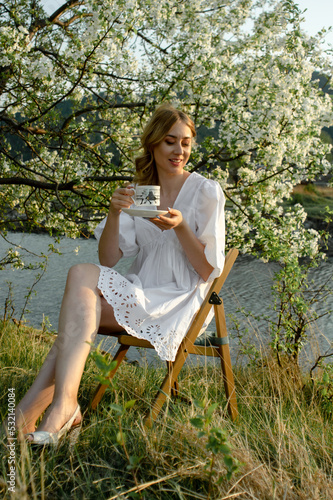 happy caucasian woman in wthite sundress with coffee cup sitting on a chair under a flowering apple tree at riverside during a sunset © DenisLubsanov
