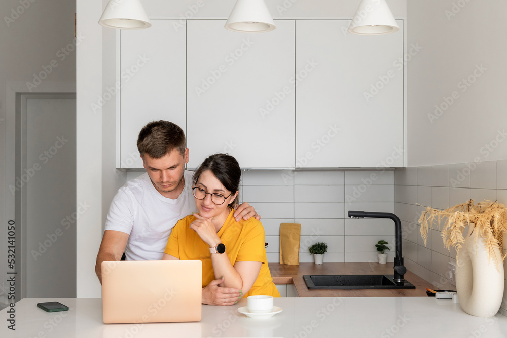Pleasant married couple is sitting in a modern kitchen looking at a laptop screen. A young family is surfing the web, shopping online or booking flights. Pleasant married couple is sitting in a modern kitchen looking at a laptop screen. A young family is surfing the web, shopping online or booking flights.
