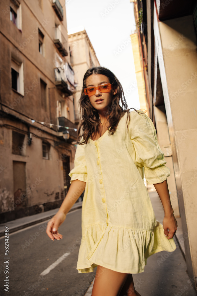Beautiful young caucasian woman posing looking at camera spending time outdoors in summer. Brunette wears sunglasses, yellow dress. Fashion trends concept