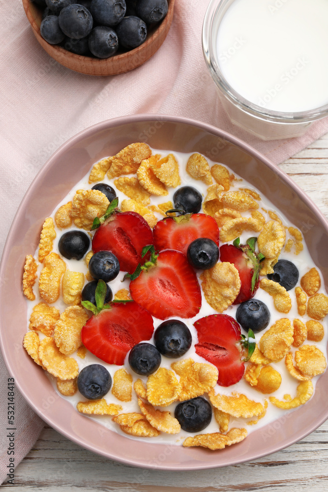 Bowl of tasty crispy corn flakes with milk and berries on white wooden table, flat lay