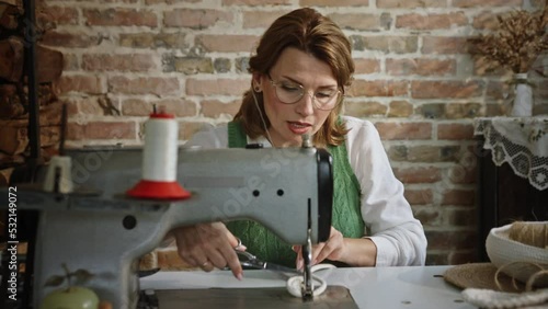 Woman sewing accessory at home. Pan around view of adult seamstress in glasses cutting off excess threads from spiral while sitting at table and creating accessory with sewing machine against brick photo