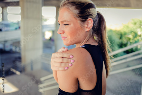 One young caucasian woman holding her sore shoulder while exercising outdoors. Female athlete suffering with painful arm injury from fractured joint and inflamed muscles during workout. © Jelena Stanojkovic