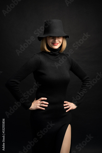 blonde woman wearing black slim dress and black hat on dark background © alipko
