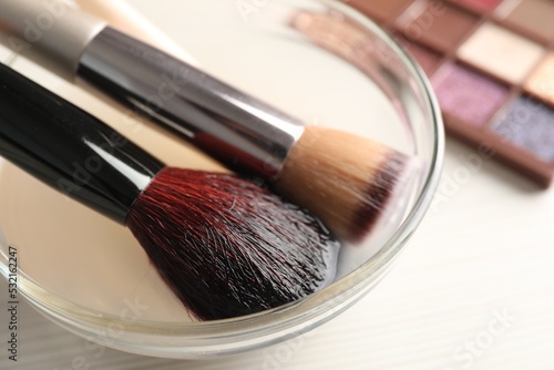 Cleaning makeup brushes in bowl with special liquid on white wooden table, closeup