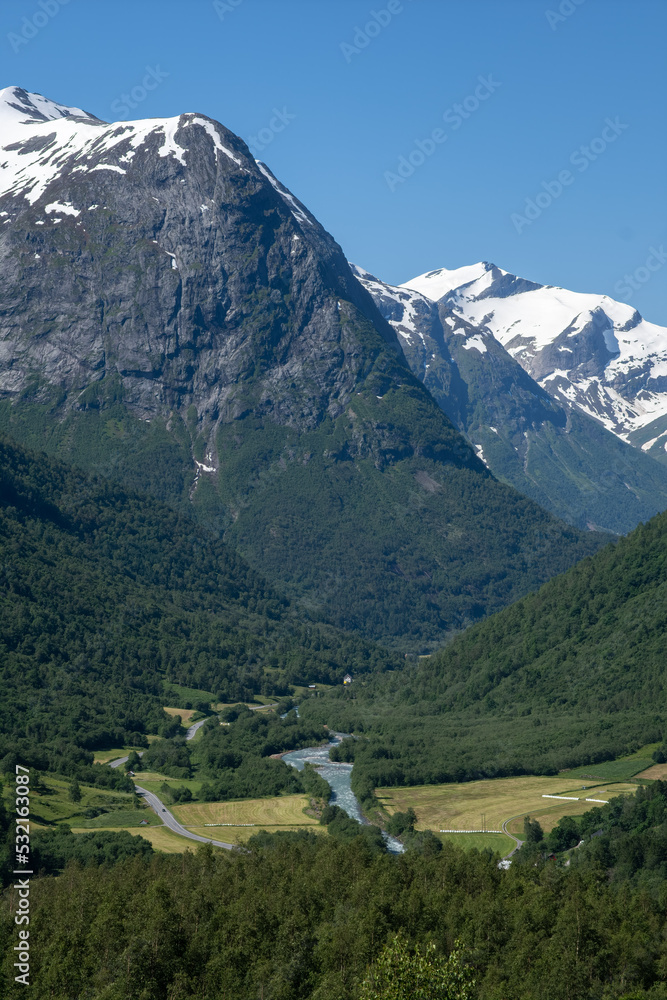 Wonderful landscapes in Norway. Vestland. Beautiful scenery of a mountain valley from the Hjelledalen lookout. Stream, snowed mountain, trees and road. Summer sunny day. Selective focus