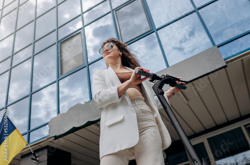 Beautiful young woman in sunglasses and white suit standing with her electric scooter near modern building and looking away