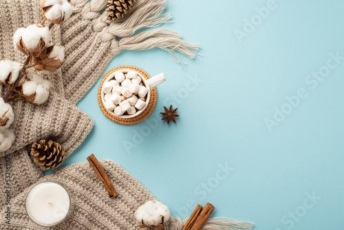 Top view photo of knitted plaid cup of hot drinking with marshmallow on rattan serving mat cotton branch candle pine cones cinnamon sticks and anise on isolated pastel blue background with copyspace