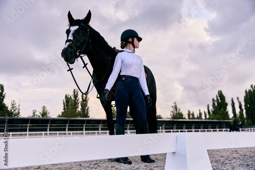 Fotografie Young sportive girl, professional jockey or horsewoman in sports uniform and helmet with black horse at riding arena