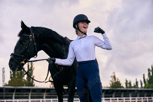 Winner. Young sportive girl, professional jockey or horsewoman in sports uniform and helmet with black horse at riding arena. Horseback riding