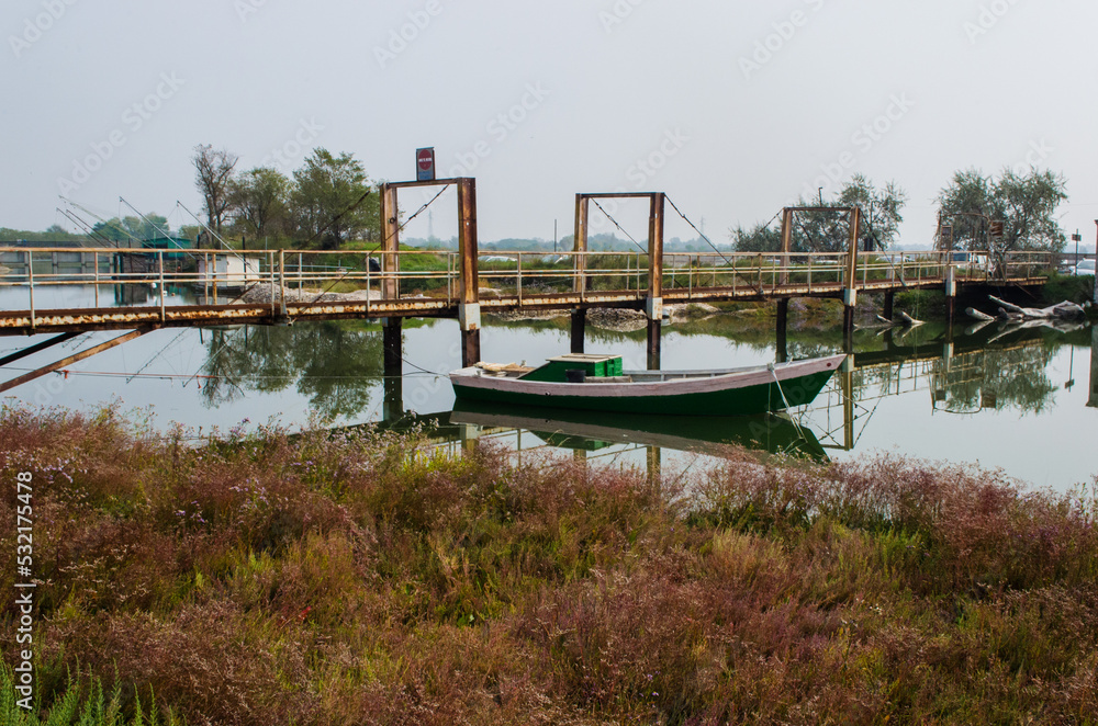 Obraz premium Una barca bianca e verde ormeggiata vicino a un pontile in un canale delle Valli di Comacchio in una giornata nuvolosa