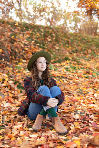 Young girl in hat and autumn clothes sit on golden fall leaves. Happy girl in green hat and plaid jacket on an autumn background. A smiling girl walking in fall park.