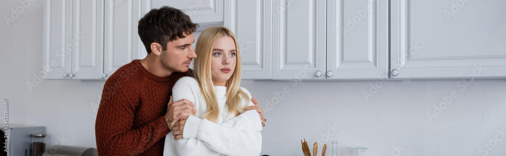 man in knitted sweater hugging blonde girlfriend in white kitchen, banner. man in knitted sweater hugging blonde girlfriend in white kitchen, banner.