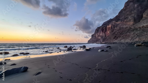 Scenic view during sunset on volcanic black sand beach Playa del Ingles in Valle Gran Rey, La Gomera, Canary Islands, Spain, Europe. Massive cliffs of the La Mercia range. Smooth waves sweeping rocks