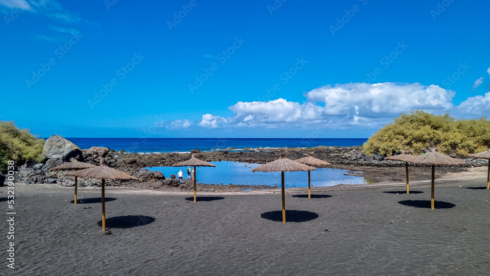Scenic sea view on beach Playa Charco del Conde without people in Valle Gran Rey, La Gomera, Canary Islands, Spain, Europe. Straw umbrellas in the dark volcanic sand. Vacation vibes in tropical bay