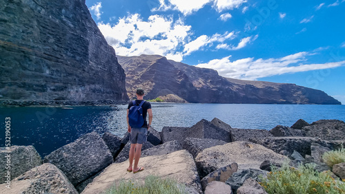 Man with backpack on scenic coastal hiking trail from Vueltas to Playas de las Arenas along massive cliffs in Valle Gran Rey on La Gomera, Canary Islands, Spain, Europe. Panoramic view from harbour