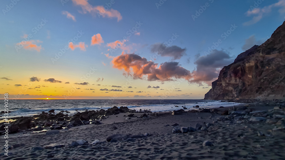 Obraz premium Scenic view during sunset on the volcanic sand beach Playa del Ingles in Valle Gran Rey, La Gomera, Canary Islands, Spain, Europe. Massive cliffs of the La Mercia range. Calm atmosphere at the seaside