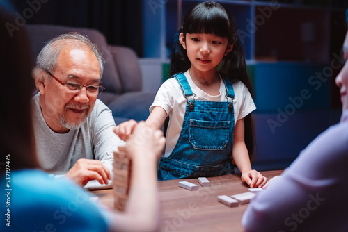 Asian happy family of little daughter play toy wooden block in home. Grandparents and young woman loving couple parent having fun play stack tower wood block game at night in living room.