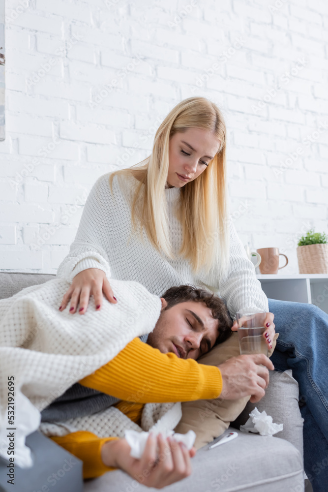 caring young woman sitting on sofa and giving glass of water to sick boyfriend.