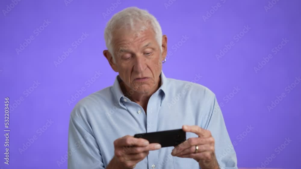 Elderly senior 70s, handsome man playing video game on phone, isolated on a purple solid background. Elderly senior 70s, handsome man playing video game on phone, isolated on a purple solid background.