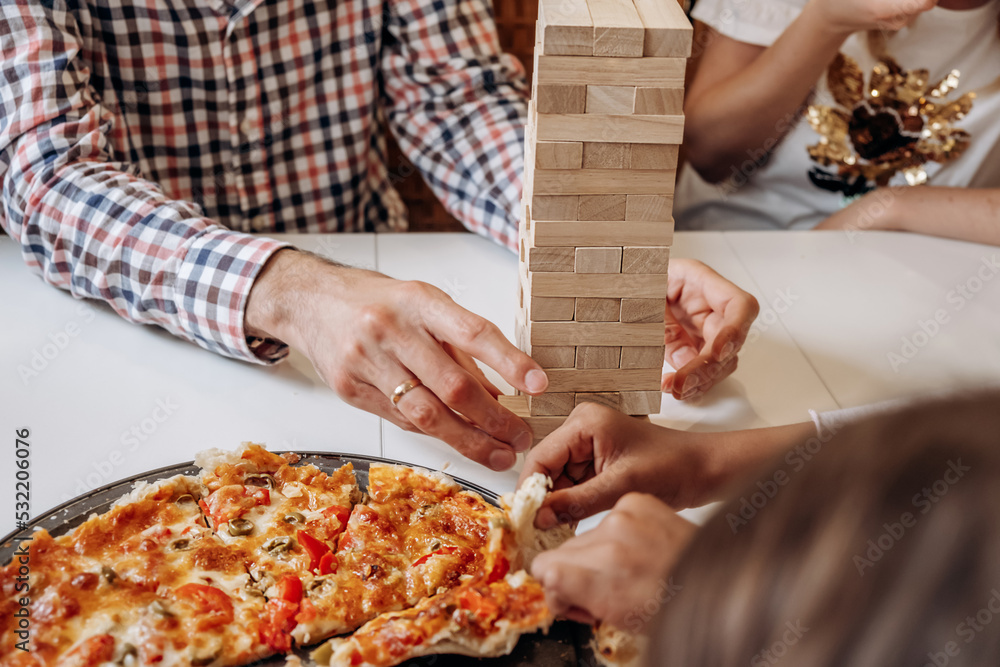 A group of multiage and multiethnic people playing jenga game on a table at home,hands close up.People eat pizza while playing.Board games concept.Time together.Stay home concept.Selective focus. A group of multiage and multiethnic people playing jenga game on a table at home,hands close up.People eat pizza while playing.Board games concept.Time together.Stay home concept.Selective focus.