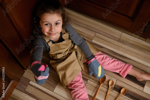 Overhead view of a baby girl, cute little baker confectioner, chef pastry in a beige apron and mittens, showing thumbs up while sitting barefoot on the kitchen floor leaning against a wooden cupboard