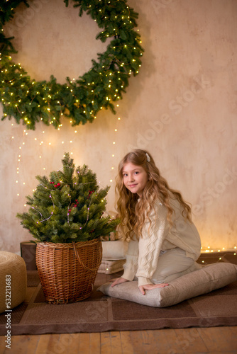 Little cute girl with blond long hair in a warm knitted sweater by a Christmas tree and a big Christmas wreath and burning garlands. Christmas mood  © capable97