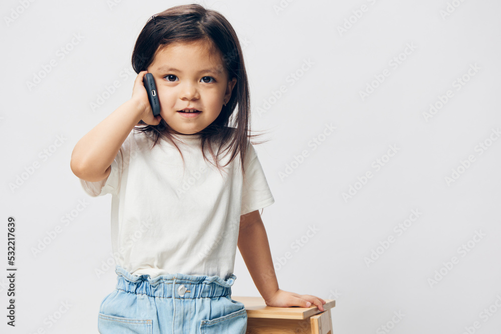 a cute little preschool girl is standing on a white background in a white T - shirt , sitting on a chair and talking on the phone looking away . The topic of children's conversation a cute little preschool girl is standing on a white background in a white T - shirt , sitting on a chair and talking on the phone looking away . The topic of children's conversation
