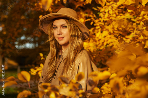 a beautiful young woman in a light hat stands near a bush with yellow autumn leaves in the sunlight