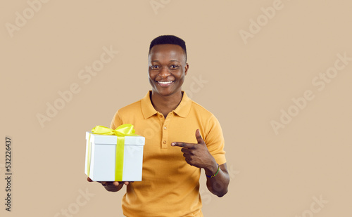 Happy handsome young man holding present tied with ribbon. Cheerful attractive African American guy in casual yellow T shirt smiling and pointing at white gift box with yellow bow in his hand photo