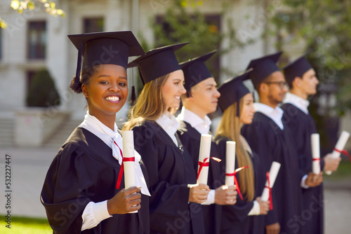 Cheerful young African American woman student in graduation gown and hat smiles during holiday in honor of end of academic year stands in line with graduates near college campus. Selective focus