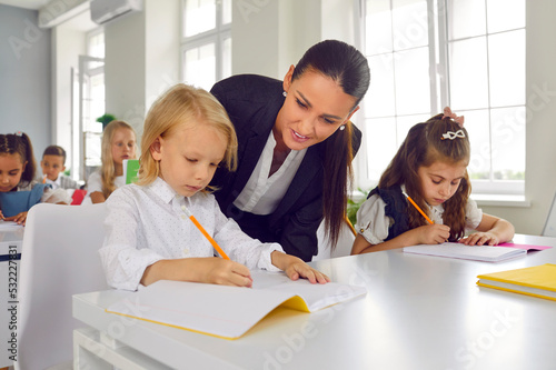 Friendly young female teacher helping little students with test tasks in primary school classroom. Teacher walks around classroom during lesson and gives hint to little schoolboy. Concept of education © Studio Romantic