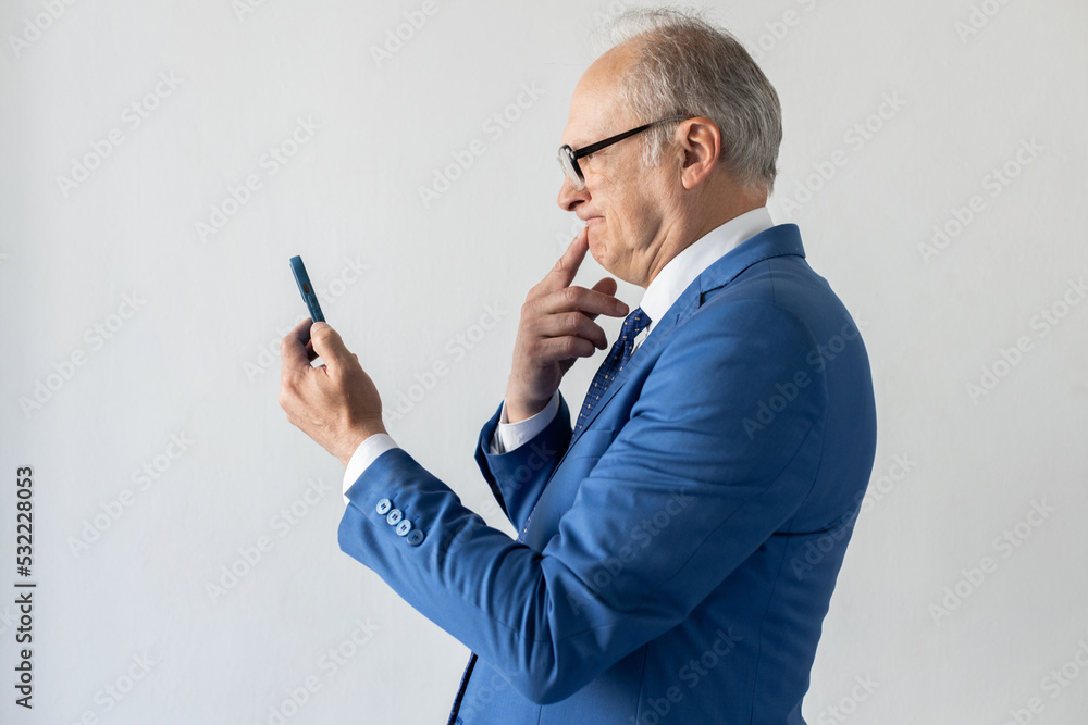 Portrait of pensive mature businessman reading message on mobile phone. Senior manager wearing formalwear and eyeglasses looking at smartphone in confusion. Mobile communication concept Portrait of pensive mature businessman reading message on mobile phone. Senior manager wearing formalwear and eyeglasses looking at smartphone in confusion. Mobile communication concept