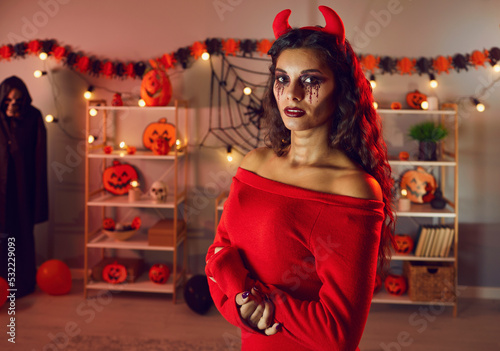 Portrait of woman in evil costume with horns, scary dark makeup celebrating Halloween indoors. Young female on ball masquerade on All Saints Hallows eve in decorated home interior. Festive. photo