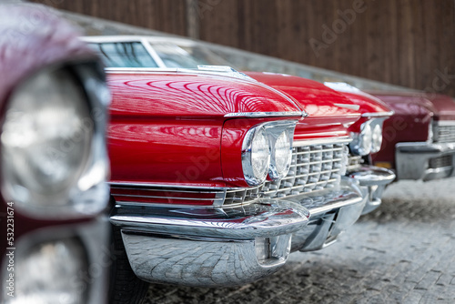 Many classic red vintage old american cars parked in row at garage, exhibition of fest. Close-up detail view beautiful retro oldtimer vehicle hood, headlight, fender and shine chrome plated bumper