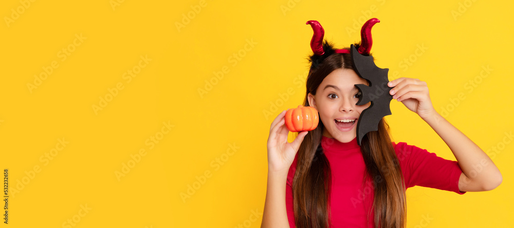amazed child in imp horns hold pumpkin and bat on yellow background, cheerfulness. Halloween kid girl portrait, horizontal poster. Banner header with copy space.
