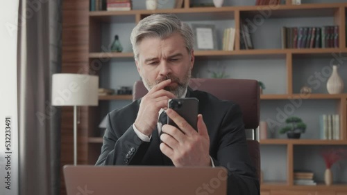 Middle-aged bearded businessman uses smartphone while sitting in office. A stylish businessman looks at the news, studies trends.