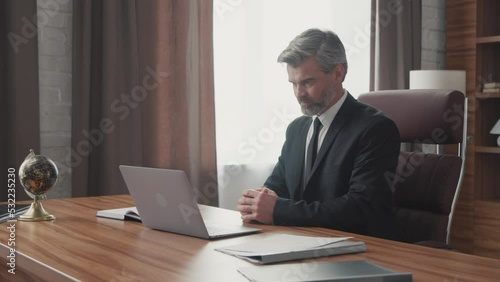 Middle aged businessman talking via online video call on his laptop while sitting at the table showing paper charts and sales statistics