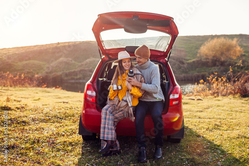Young couple in love relaxing in car trunk by autumn lake drinking tea from vacuum flask. Man and woman travel by auto.