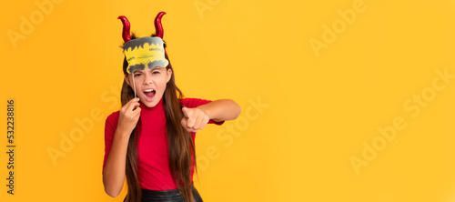 devil kid with pumpkin. trick or treat. teen girl point finger on yellow background. Halloween kid girl portrait, horizontal poster. Banner header with copy space.