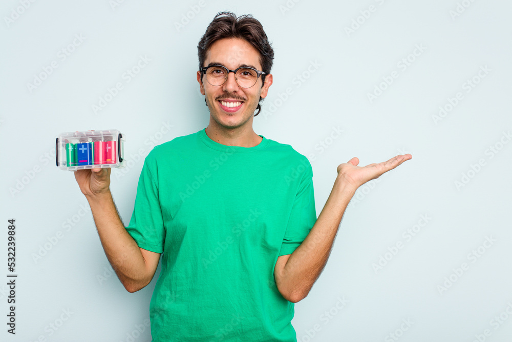 Young hispanic man holding battery box isolated on white background showing a copy space on a palm and holding another hand on waist. Young hispanic man holding battery box isolated on white background showing a copy space on a palm and holding another hand on waist.
