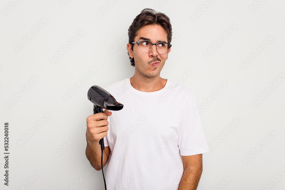 Young hispanic man holding a hairdryer isolated on white background confused, feels doubtful and unsure. Young hispanic man holding a hairdryer isolated on white background confused, feels doubtful and unsure.