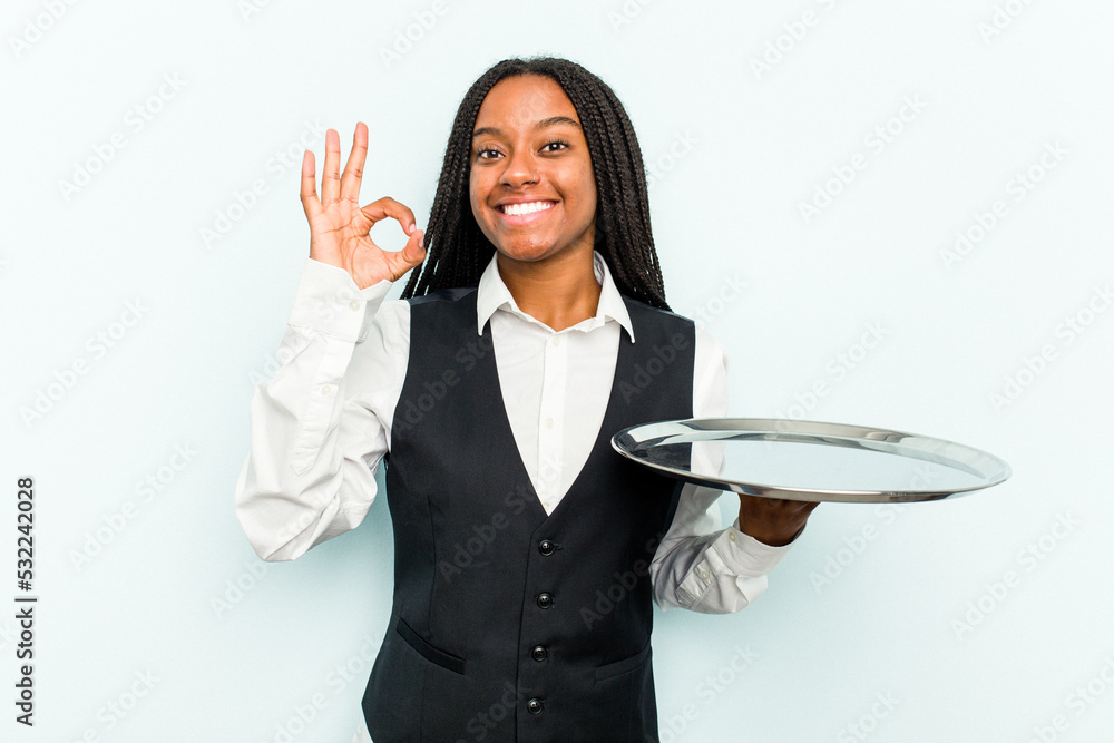 Young African American waitress woman holding a tray isolated on blue background cheerful and confident showing ok gesture. Young African American waitress woman holding a tray isolated on blue background cheerful and confident showing ok gesture.
