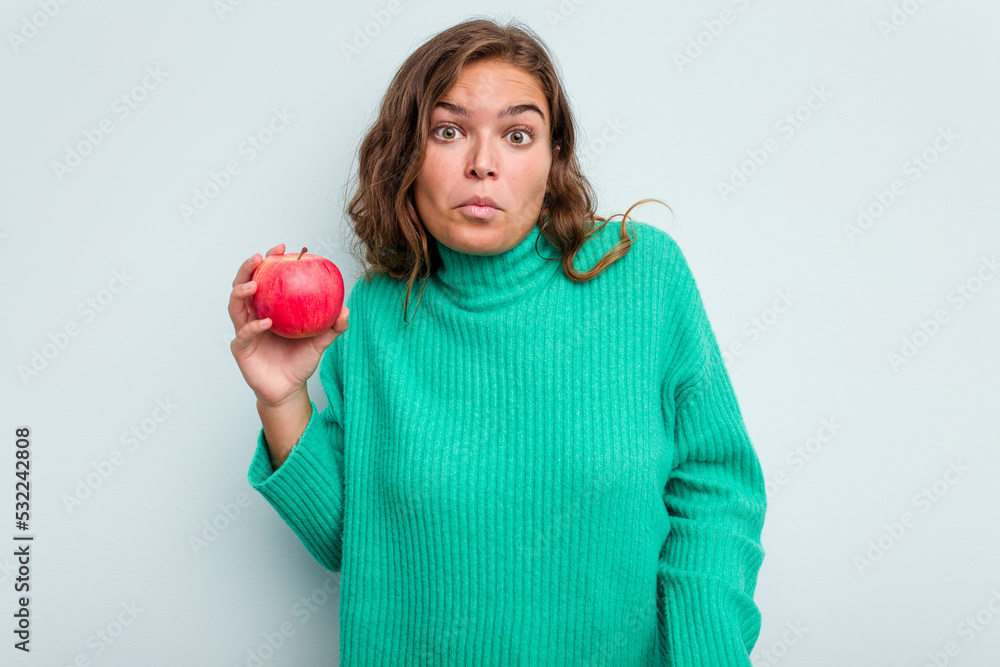 Young caucasian woman with an apple isolated on blue background shrugs shoulders and open eyes confused. Young caucasian woman with an apple isolated on blue background shrugs shoulders and open eyes confused.