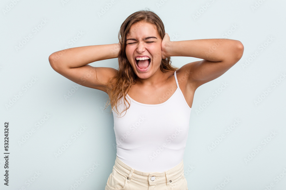 Young caucasian woman isolated on blue background covering ears with hands trying not to hear too loud sound. Young caucasian woman isolated on blue background covering ears with hands trying not to hear too loud sound.