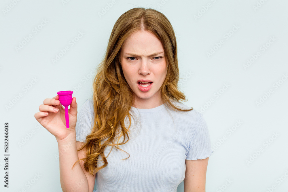 Young caucasian woman holding menstrual cup isolated on blue background screaming very angry and aggressive. Young caucasian woman holding menstrual cup isolated on blue background screaming very angry and aggressive.