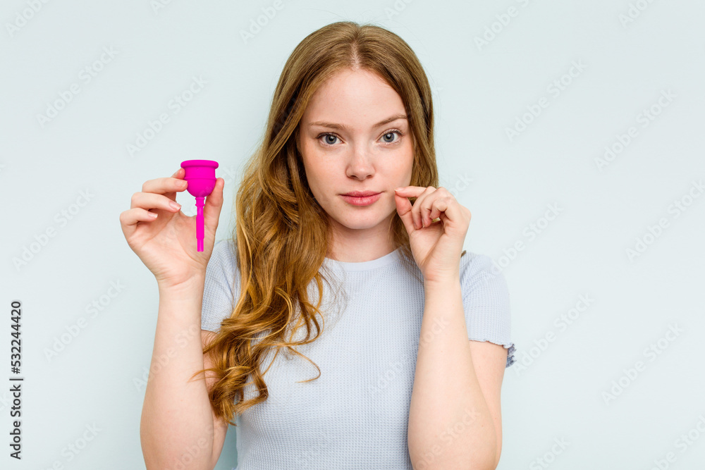 Young caucasian woman holding menstrual cup isolated on blue background with fingers on lips keeping a secret. Young caucasian woman holding menstrual cup isolated on blue background with fingers on lips keeping a secret.