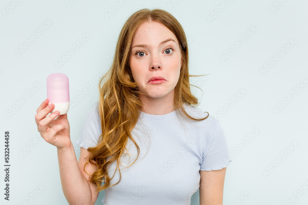 Young caucasian woman holding deodorant isolated on blue background shrugs shoulders and open eyes confused. Young caucasian woman holding deodorant isolated on blue background shrugs shoulders and open eyes confused.