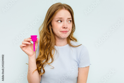 Young caucasian woman holding menstrual cup isolated on blue background confused, feels doubtful and unsure.